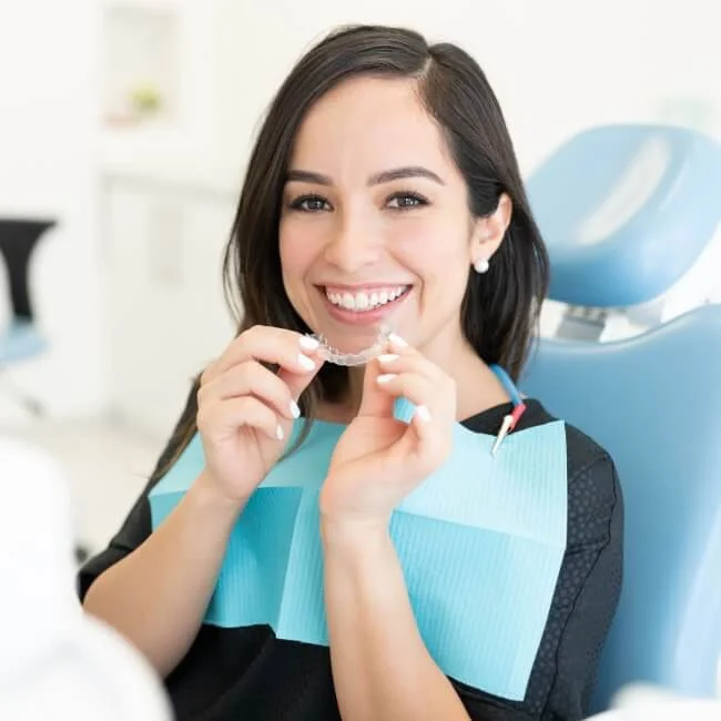 Woman smiling while holding SureSmile® clear aligners in front of her mouth in a dentist chair