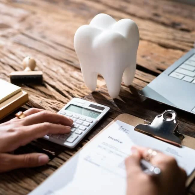 One hand on a calculator while another hand holds a pen over a clipboard filling out a dental insurance form on a wooden desk with a tooth sculpture on it