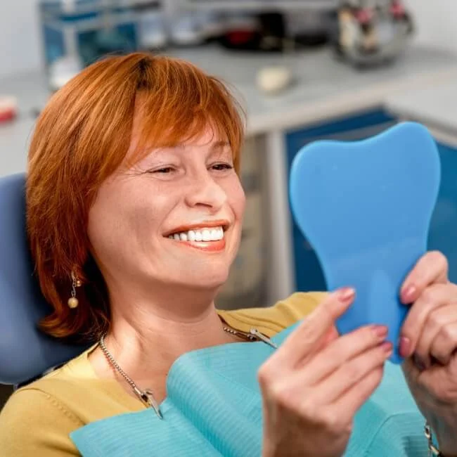 Red-haired woman sitting in a dentist chair smiling at a tooth-shaped mirror