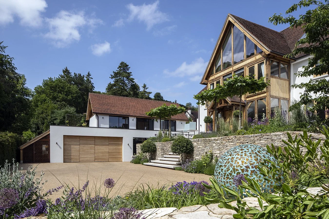 Modern house with a wooden and glass design, surrounded by lush greenery and plants, under a blue sky with clouds.