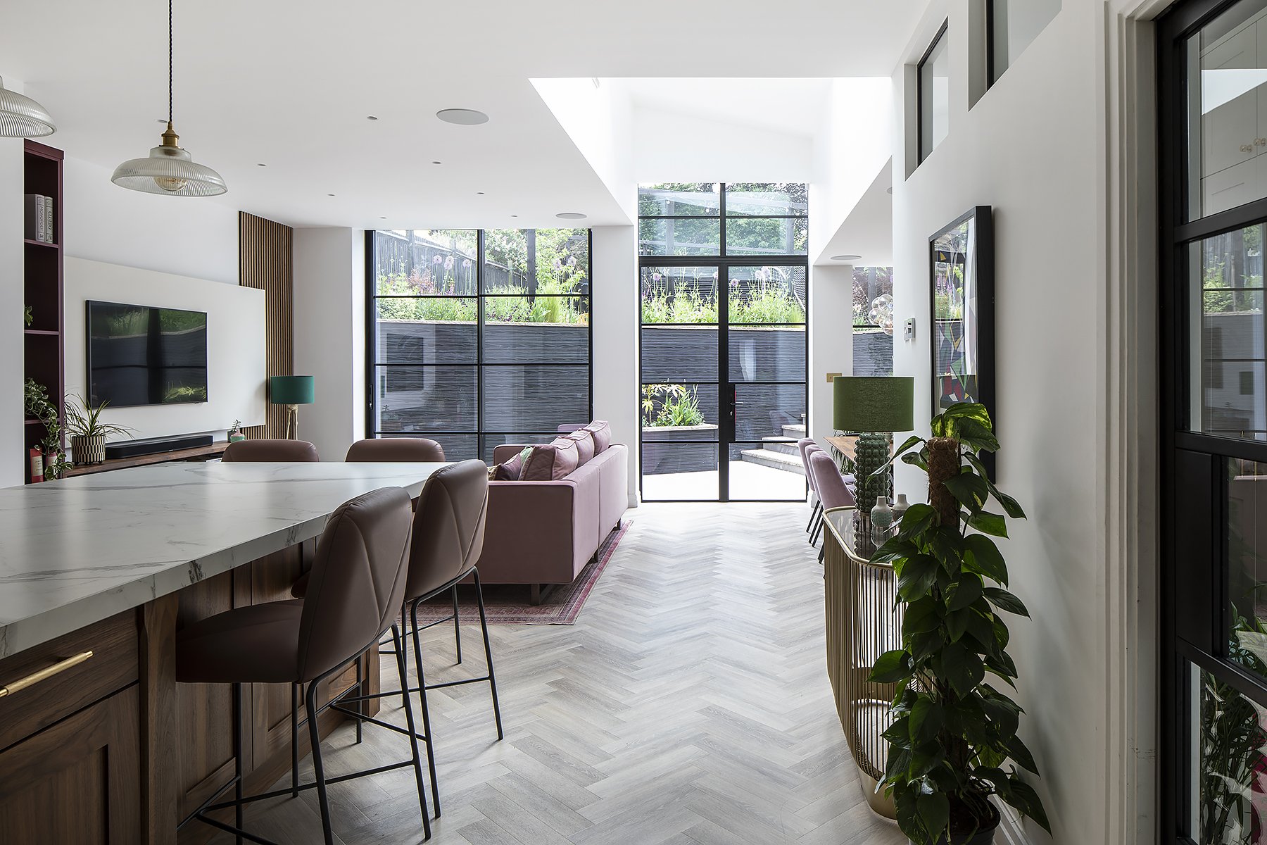 Modern living room with large windows, pink sofa, and greenery outside. Kitchen area with marble countertop and bar stools. Decorative plants and artwork.