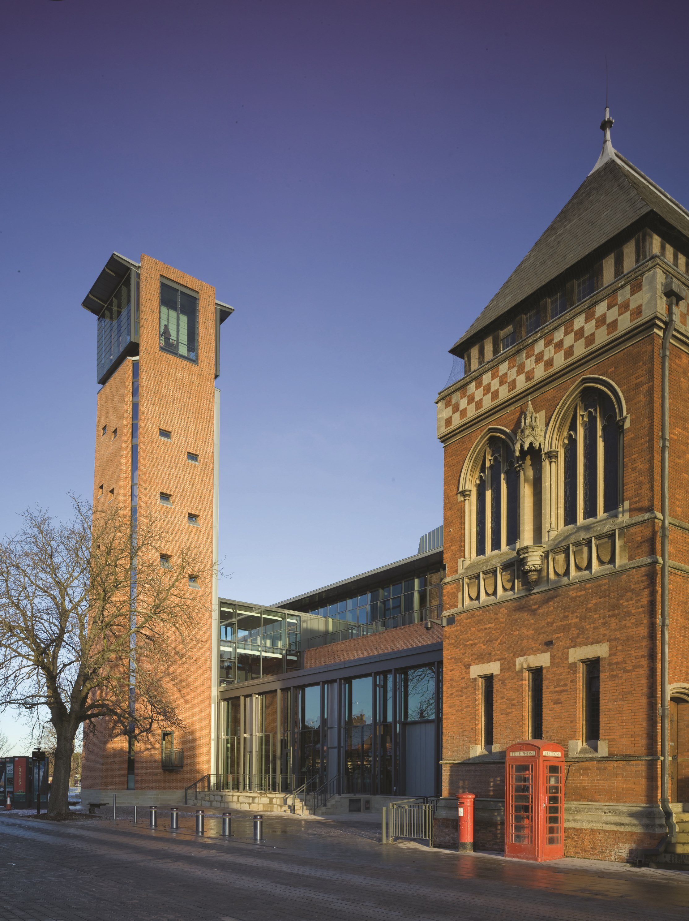 A historic red-brick building with gothic arches and a tower, connected to a modern glass extension, with a red telephone booth and leafless tree in the foreground.
