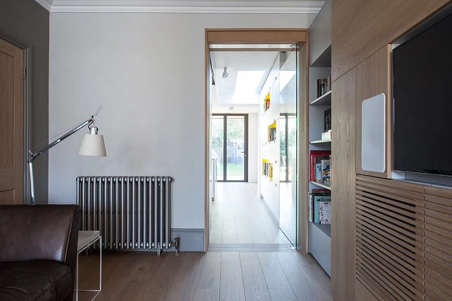 Interior view of a modern house with a hallway leading to a bright outdoor area, featuring a radiator, a reading chair, a floor lamp, and built-in shelves with books.