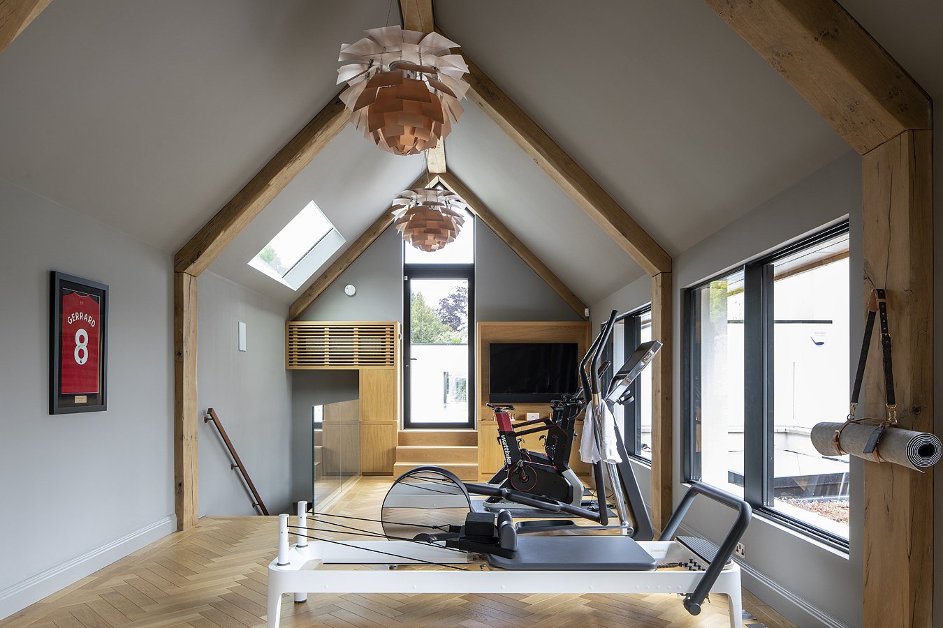 Home gym with two exercise machines, large windows, skylights, wooden beams, hanging lamps, a wall-mounted TV, and a framed sports jersey.