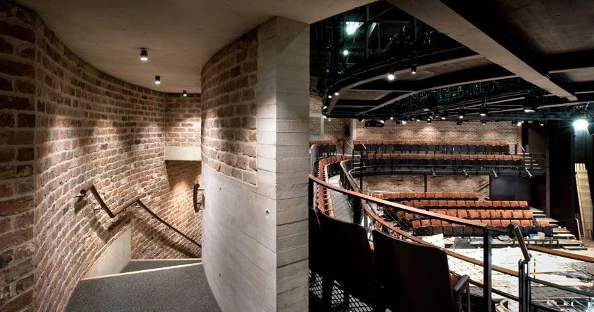 The image shows the interior of a theater with a stage, seating area, and concrete walls with brick accents.