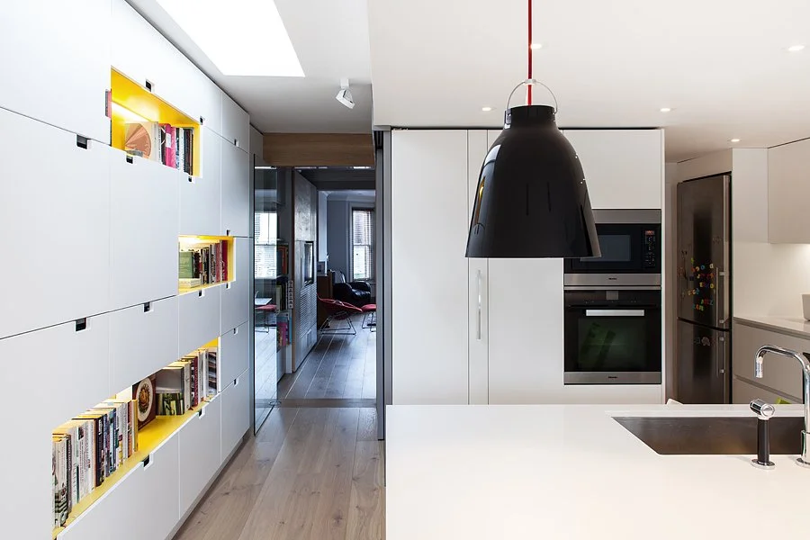Modern kitchen with white cabinetry, black pendant light, built-in oven, microwave, and refrigerator, with a view into a living area with chairs and windows.