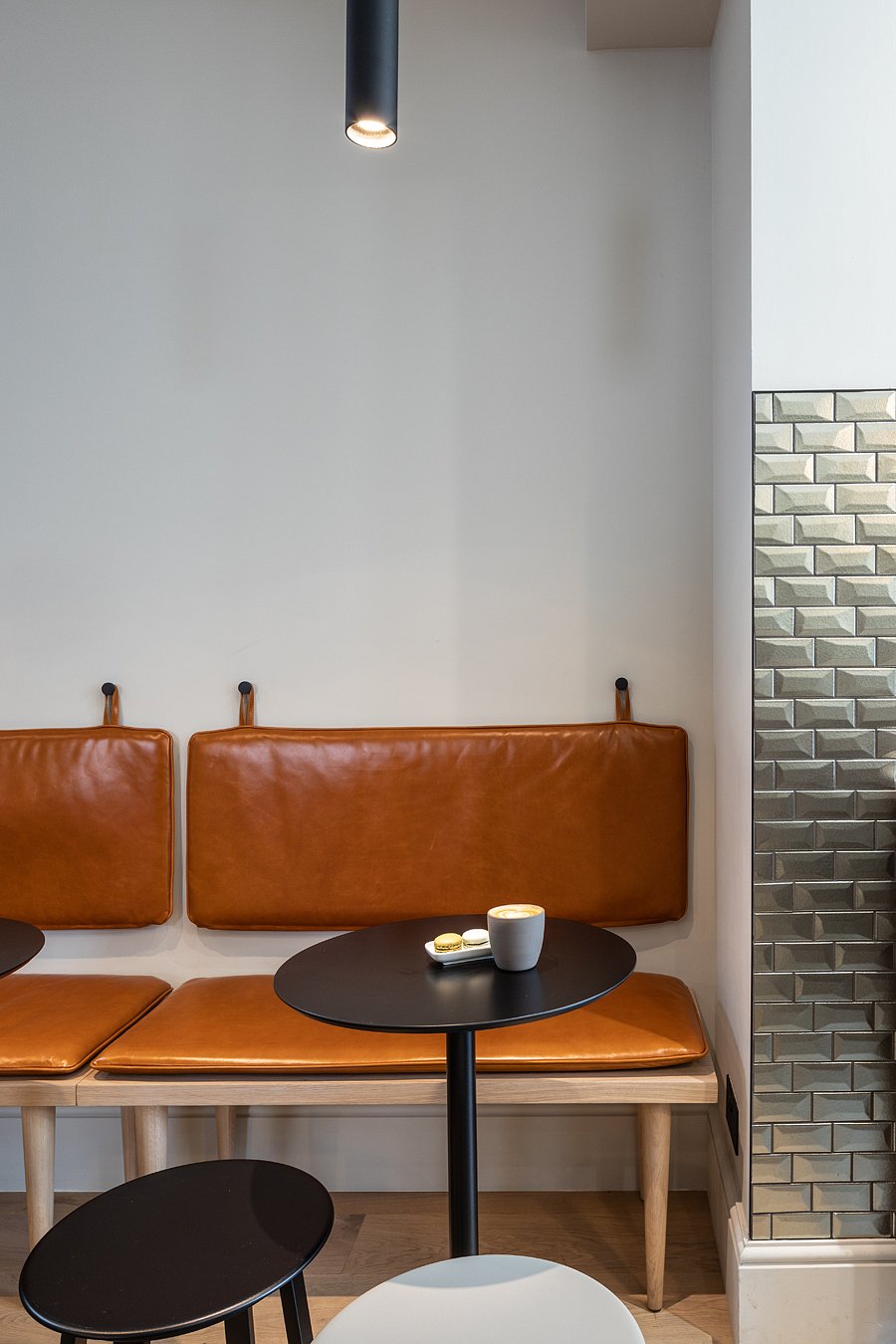 Interior of a cafe featuring a brown leather bench, small black round tables, a modern light fixture, and a textured gray brick wall on the right.