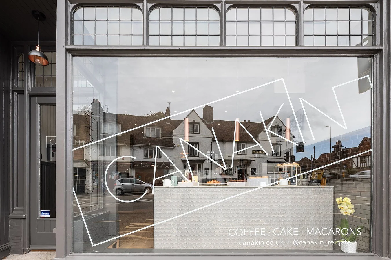 The window of a cafe named 'CANAKIN' with white geometric design and text indicating they serve coffee, cake, and macarons. The interior shows a white counter with some items, a potted plant with yellow flowers outside, and reflections of residential