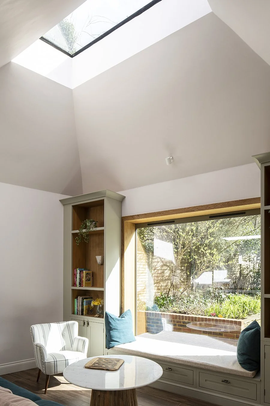 Living room with a large window seat, bookshelf, and armchair, illuminated by natural light from a skylight, overlooking a garden.
