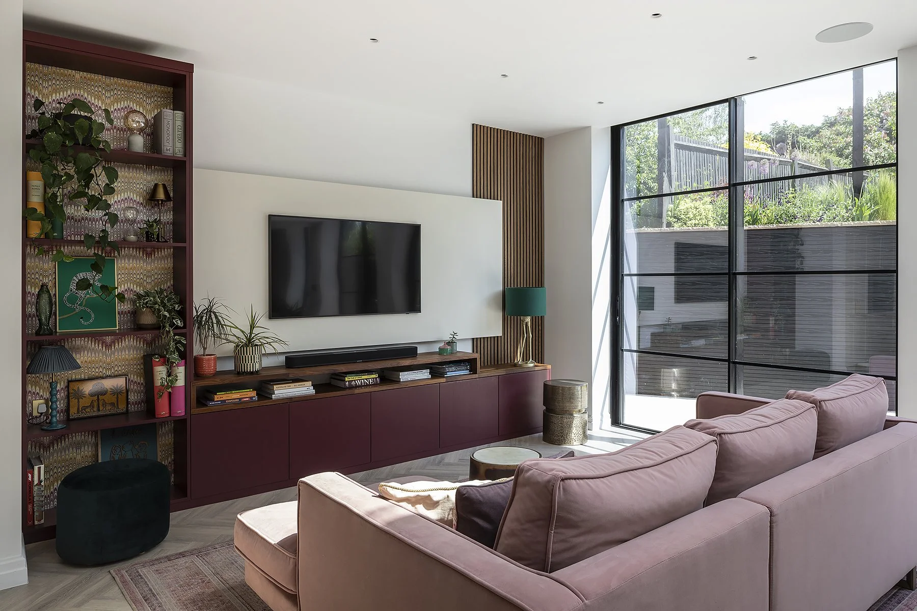 Modern living room with large floor-to-ceiling window, pink sectional sofa, TV mounted on white wall, colorful bookshelf, and decorative items.