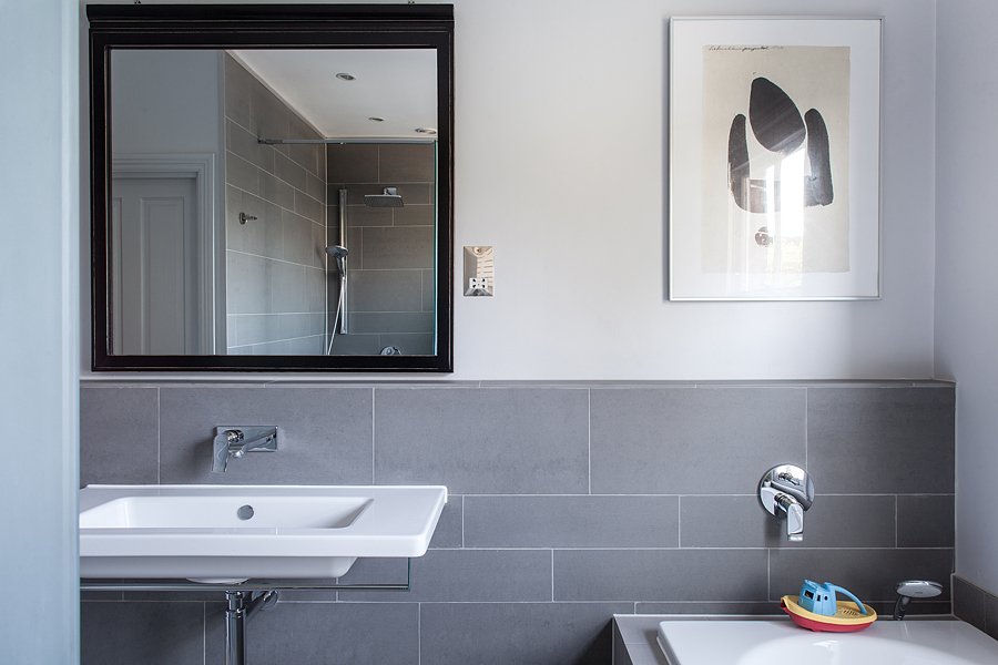 Modern bathroom with white sink and gray tiled walls, a framed abstract picture, a bathroom mirror reflecting a shower, and a toy boat on the bathtub.