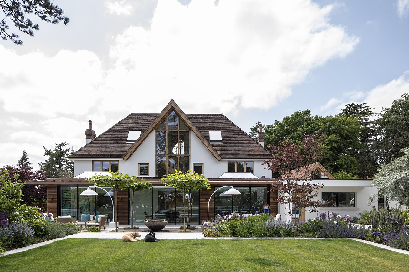 A modern house with a gable roof, large glass windows, and a front yard with green grass, flowering plants, and outdoor furniture, under a partly cloudy sky.