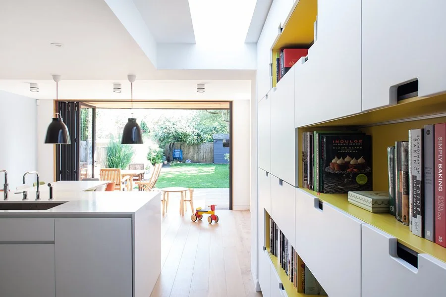 Modern, open-concept kitchen and dining area with white cabinetry, a large window looking to a backyard with grass, trees, and outdoor furniture, and a small red and yellow toy on the light wood floor.