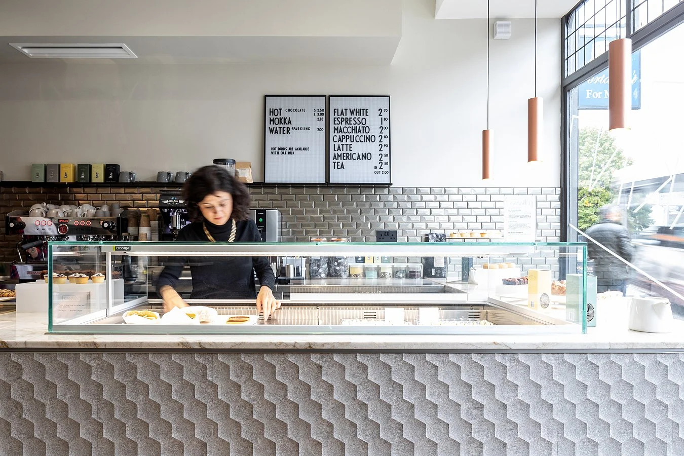 A woman behind a coffee shop counter with a display case of pastries, coffee equipment, and a menu board in the background.