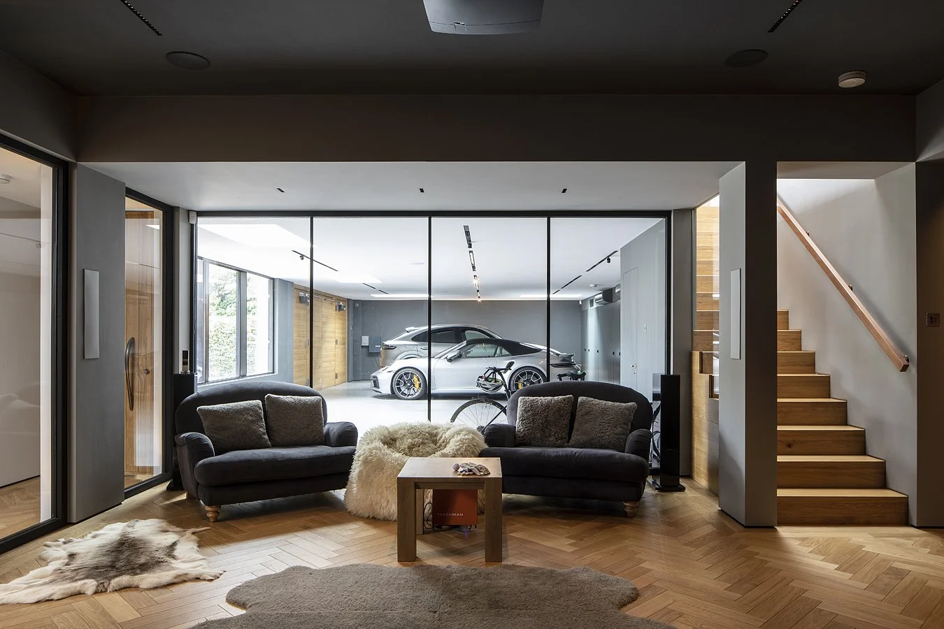 Modern living room with two black sofas, a furry throw, and a small wooden table in front. Large glass wall shows a garage with two luxury cars and a bicycle. Wooden floors and stairs on the right lead upstairs.