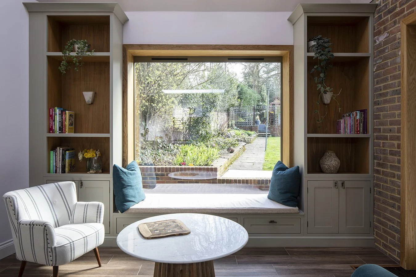 Interior of a cozy living room with a large window seat, blue pillows, built-in shelves with books and decorations, a striped armchair, and a round white coffee table, overlooking a lush garden outside.