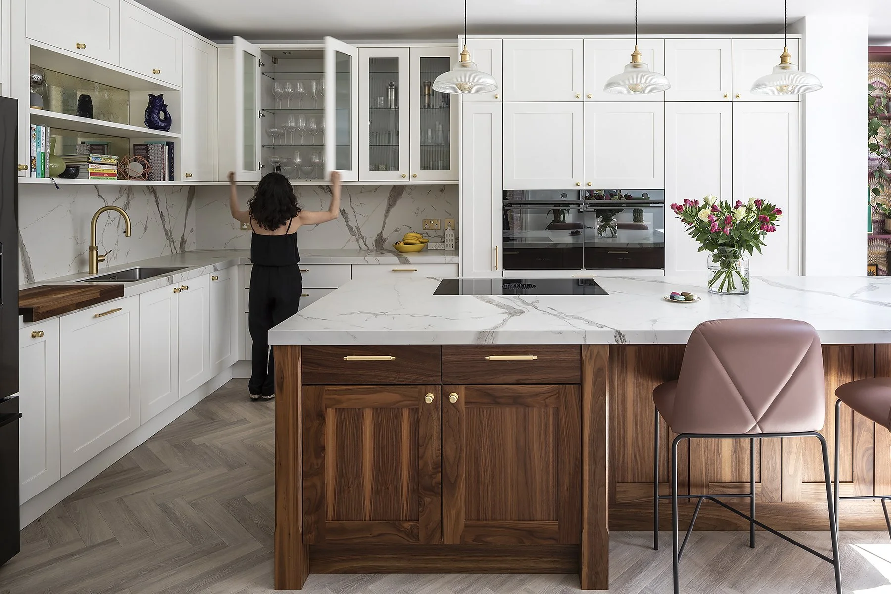 Modern kitchen with white cabinetry, marble countertops, a kitchen island with wood finish, and pendant lights. A woman is opening upper cabinets, and a vase with pink flowers is on the island.