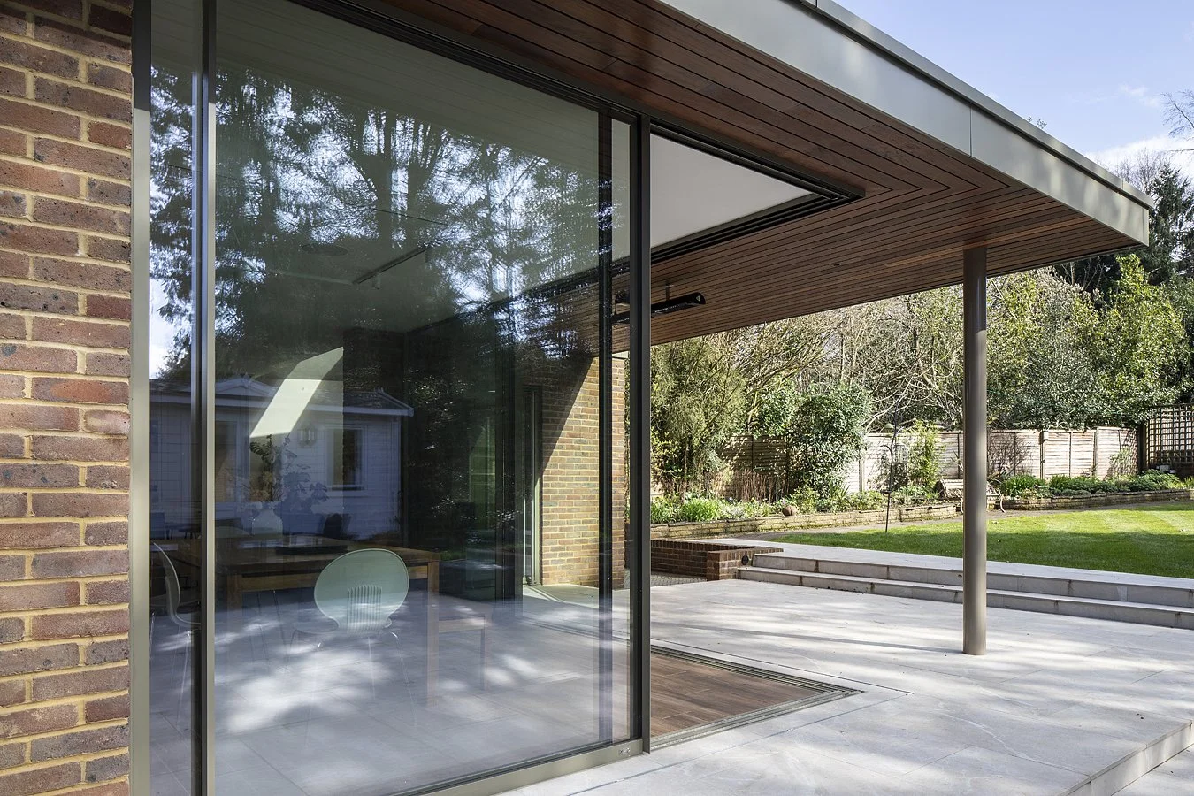 Modern house exterior with large glass sliding doors, brick wall, wooden ceiling, metal support post, and a spacious backyard with grass, trees, and landscaped garden.