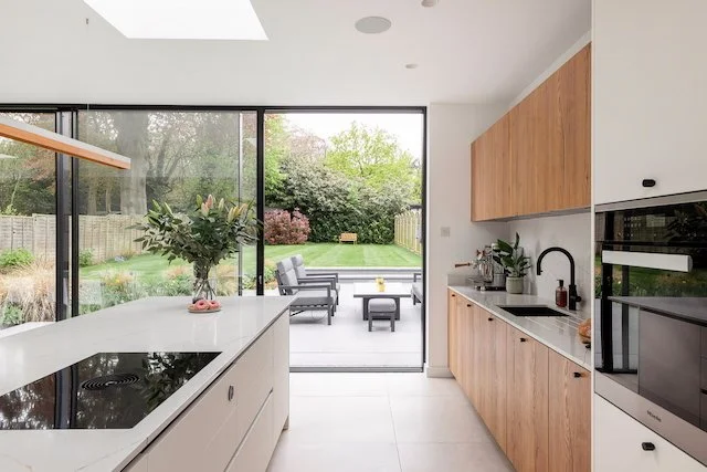 Modern kitchen with white countertops, wooden cabinets, black sink and faucet, and large glass sliding door leading to a garden outdoor patio with seating and a lawn.