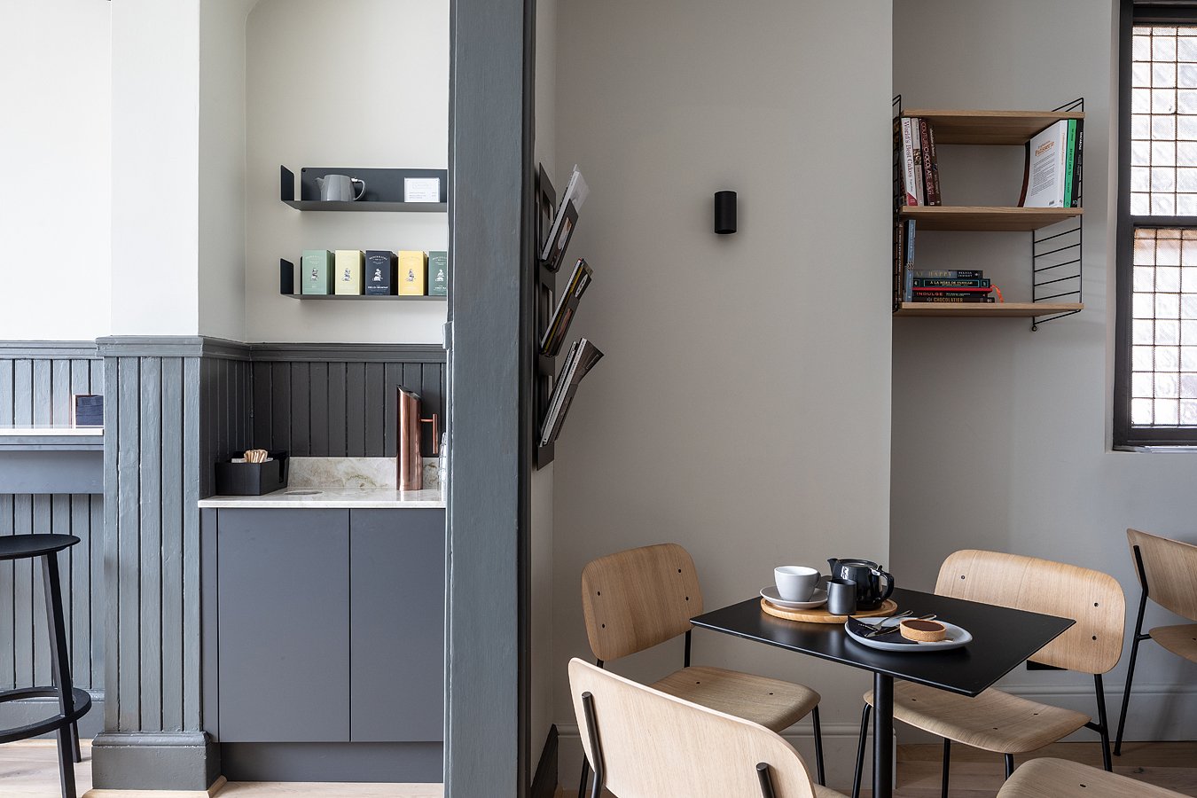 A cozy cafe corner with a small black table set for tea, surrounded by light wooden chairs, with shelves of books and kitchenware on the walls, natural light coming through a window.
