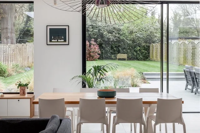 Dining area with a wooden table and six white chairs, large glass sliding doors leading to a garden with green grass, bushes, trees, and a wooden fence, and a modern hanging light fixture.