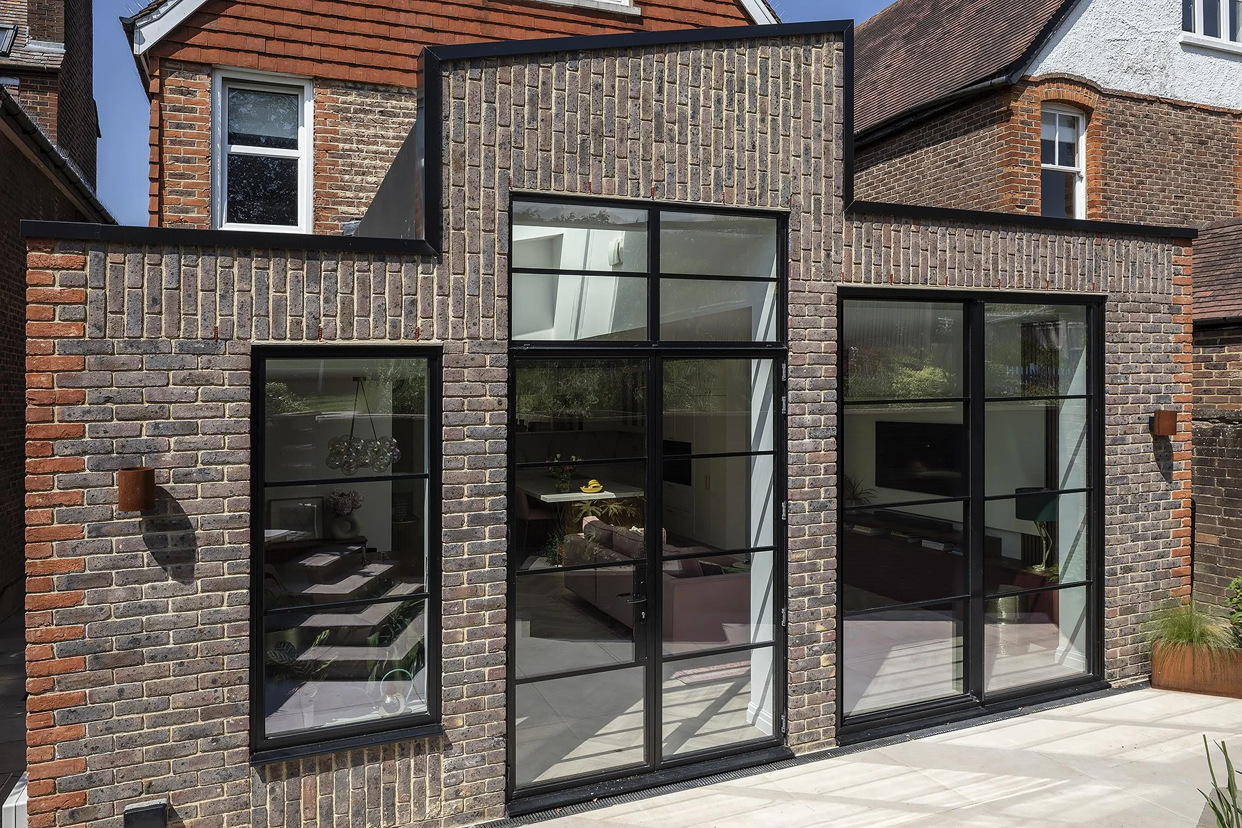 Modern brick house with large black-framed glass doors and windows, showing a living room and dining area inside.