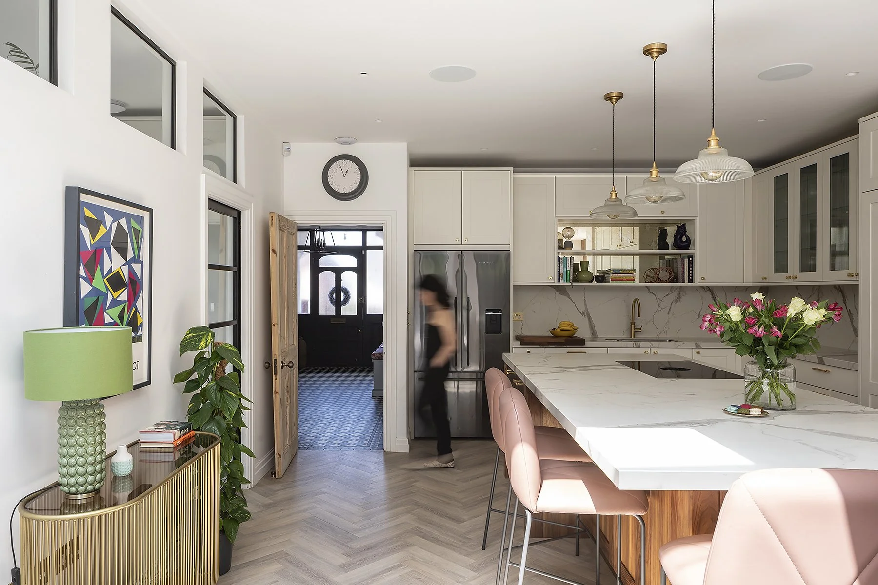 Modern kitchen with white cabinets, marble countertops, pink bar stools, and a vase of pink and white flowers on the island. A woman is walking past a stainless steel refrigerator.