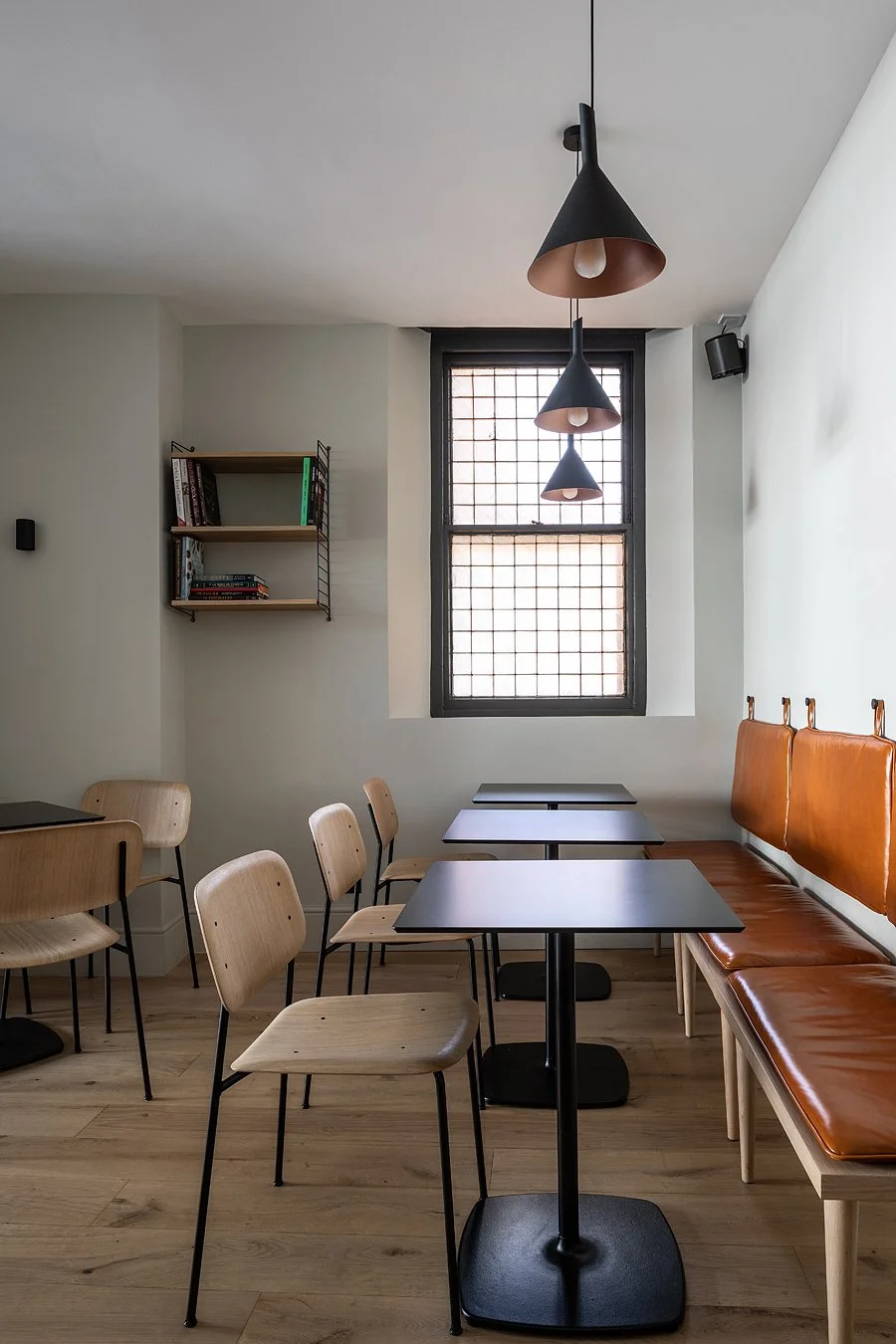 Interior of a modern cafe featuring wooden chairs, black tables, a leather-upholstered bench, a window with grid pattern, three black cone pendant lights, a small bookshelf, and wooden flooring.