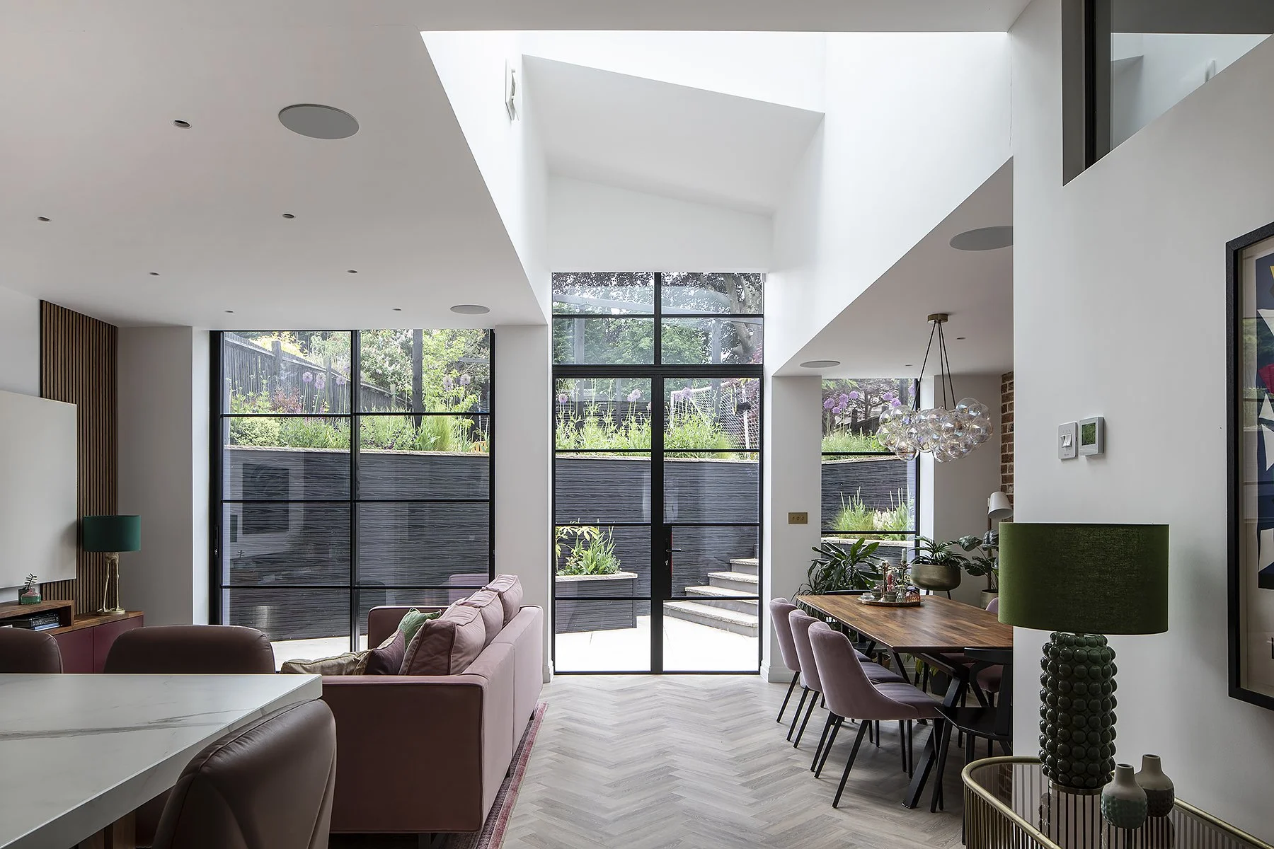 Modern living and dining room with large windows and sliding glass door, pink and leather chairs, wooden dining table, globe pendant light, green table lamp, and indoor plants.