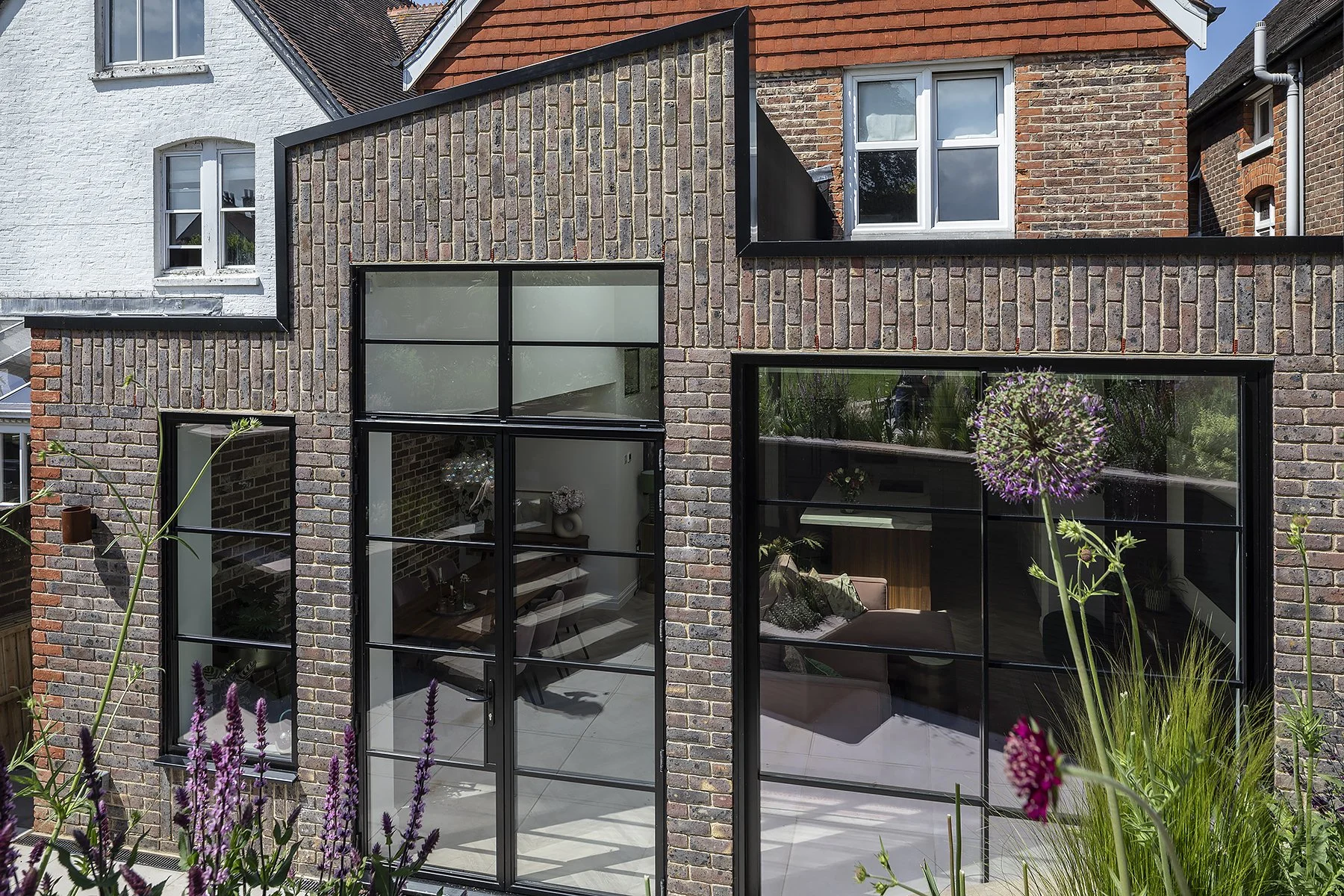Modern brick house with large black-framed glass windows and garden plants in the foreground.