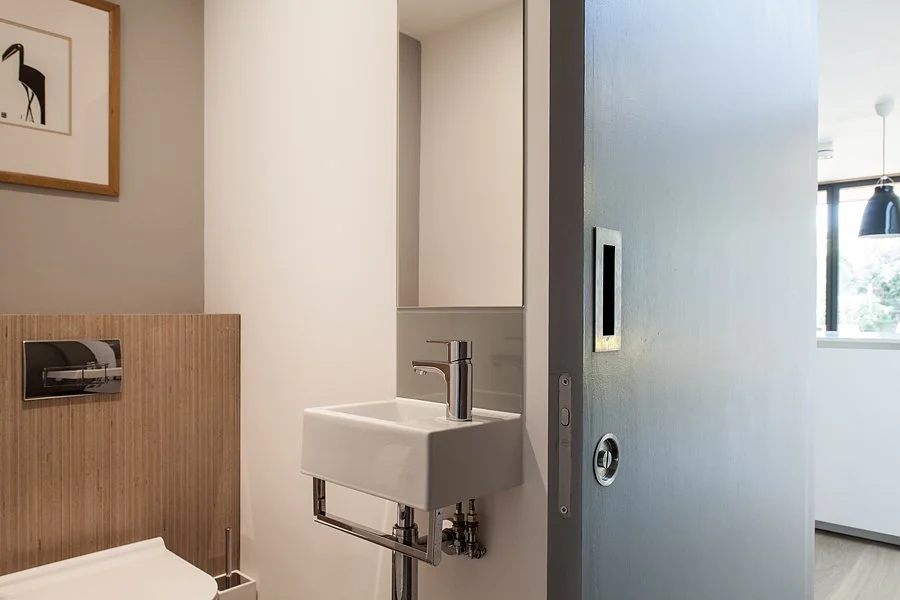 Small bathroom with a white sink, a mirror cabinet, a wooden wall behind the toilet, a picture of a zebra on the gray wall, and a partially open sliding door leading to a brightly lit room with large windows.