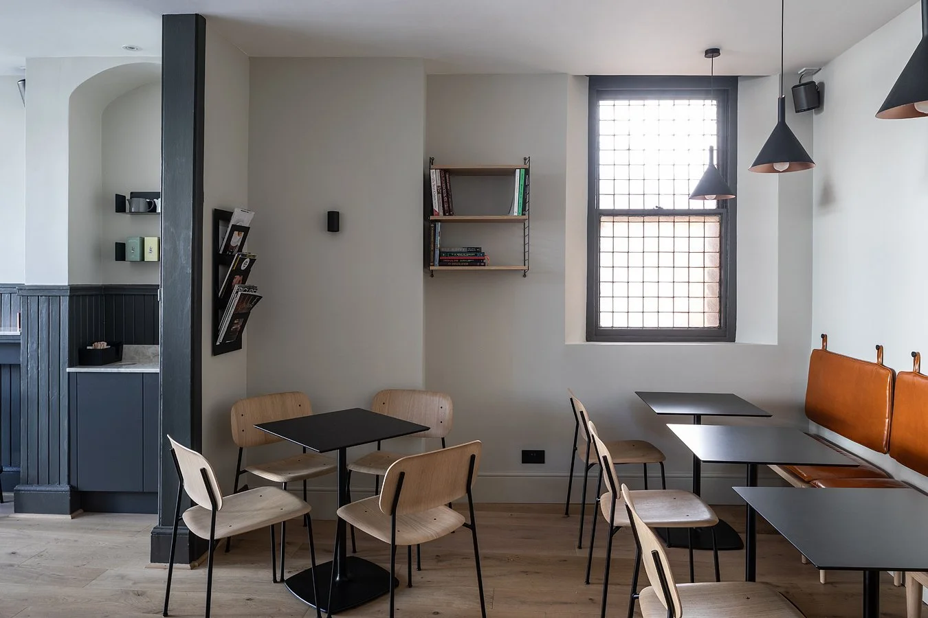 Interior of a modern cafe with black tables, wooden chairs, and a cushioned bench against the wall, with pendant lights and a window with black grid.