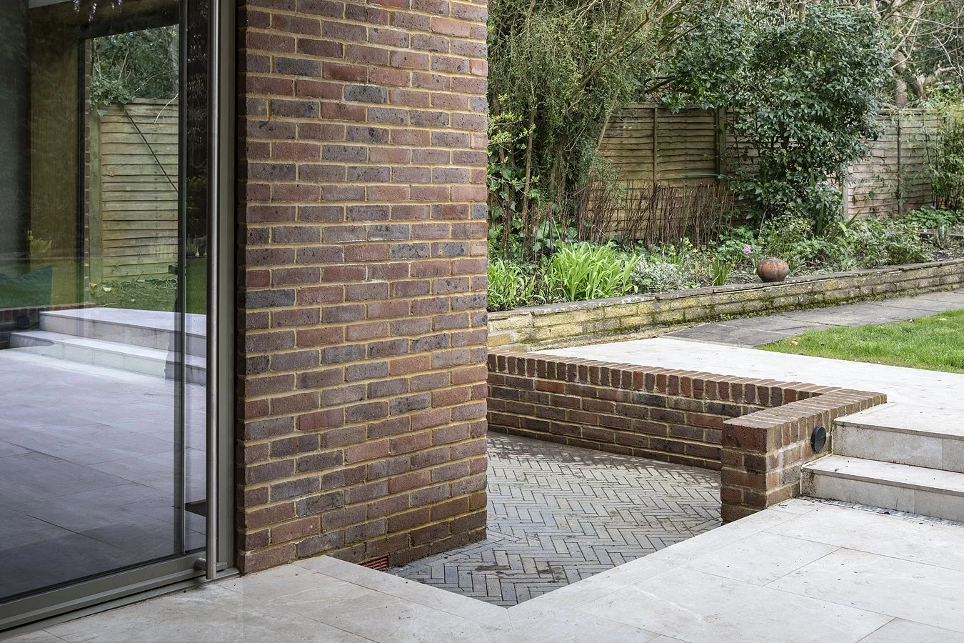 View of a backyard garden with brick wall, paved patio, and greenery, seen from a house patio with glass sliding doors.