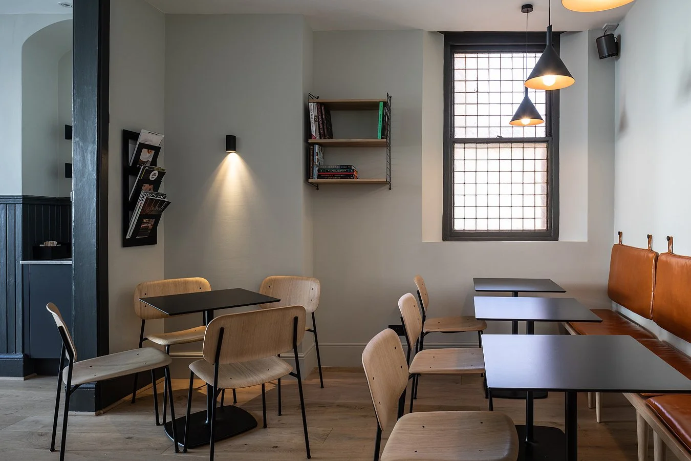 A cozy cafe interior with wooden chairs, black tables, a bench with brown leather cushions, a window with black grid pattern, hanging black pendant lights, and minimalist wall decor.