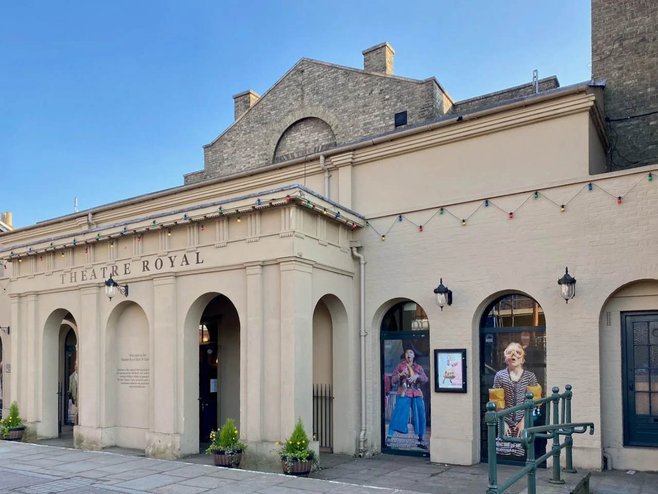 Exterior of the Theatre Royal building with string lights, posters, and potted plants outside, under a clear blue sky.