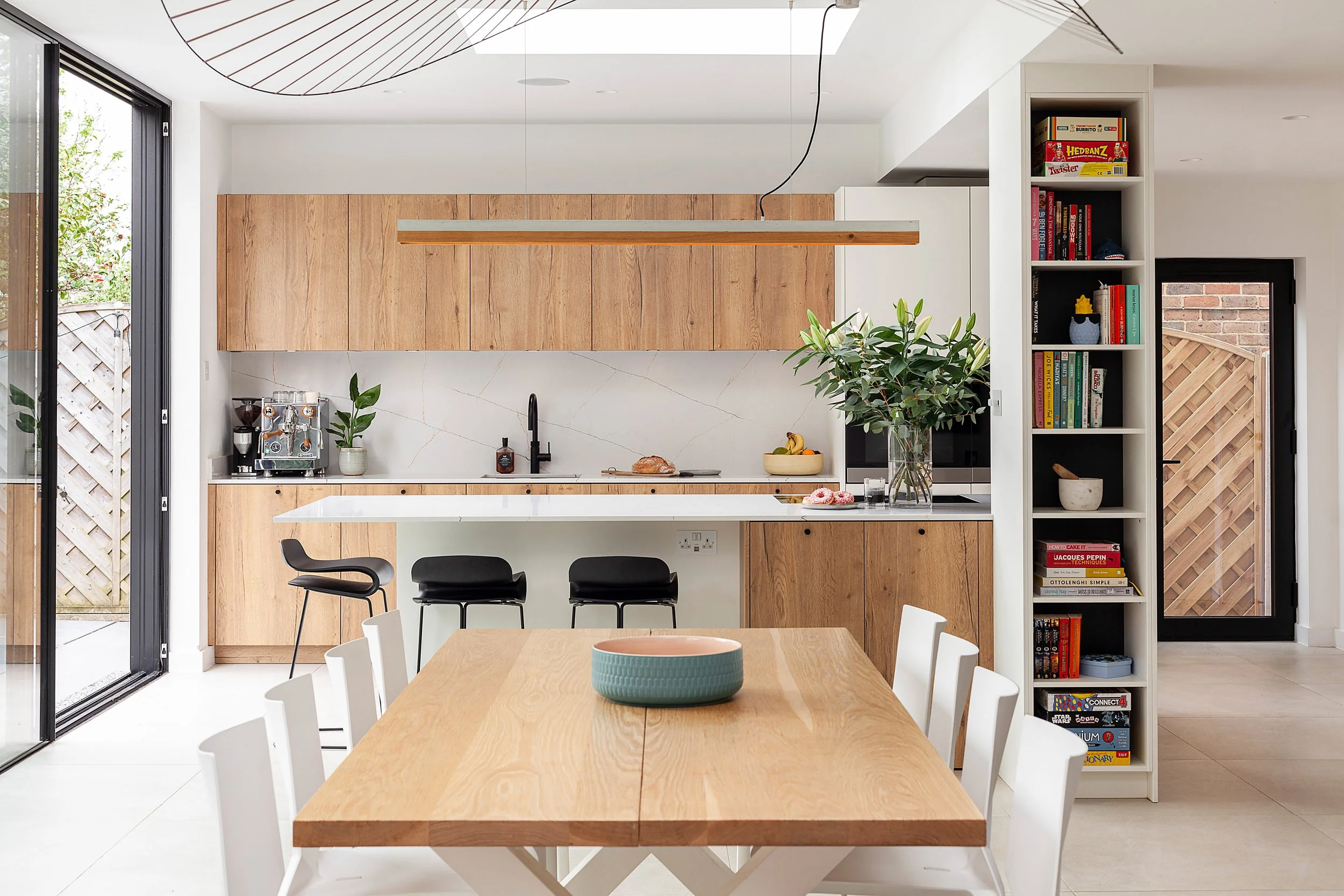 Modern kitchen with wooden cabinets, white countertops, and a dining table with white chairs. There is a large vase with greenery on the counter, a bookshelf with books, and a sliding glass door leading outside.