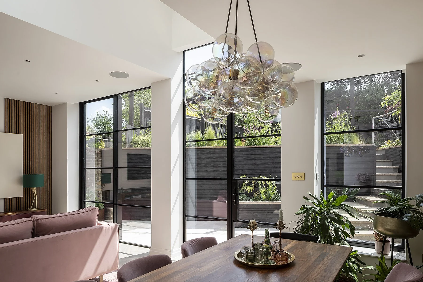 Modern dining room with large floor-to-ceiling windows, a wooden dining table with candles and glasses, lush green plants, a pink sofa, and a contemporary chandelier hanging from the ceiling.