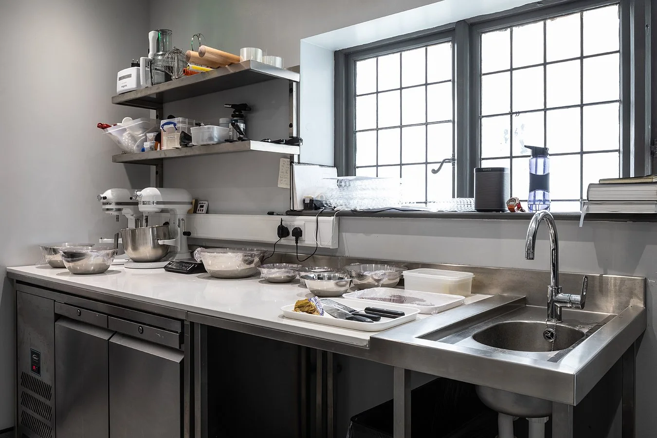 A commercial kitchen workspace with stainless steel counter, open shelving holding kitchen utensils and containers, and a large window with a grid pattern letting in natural light.