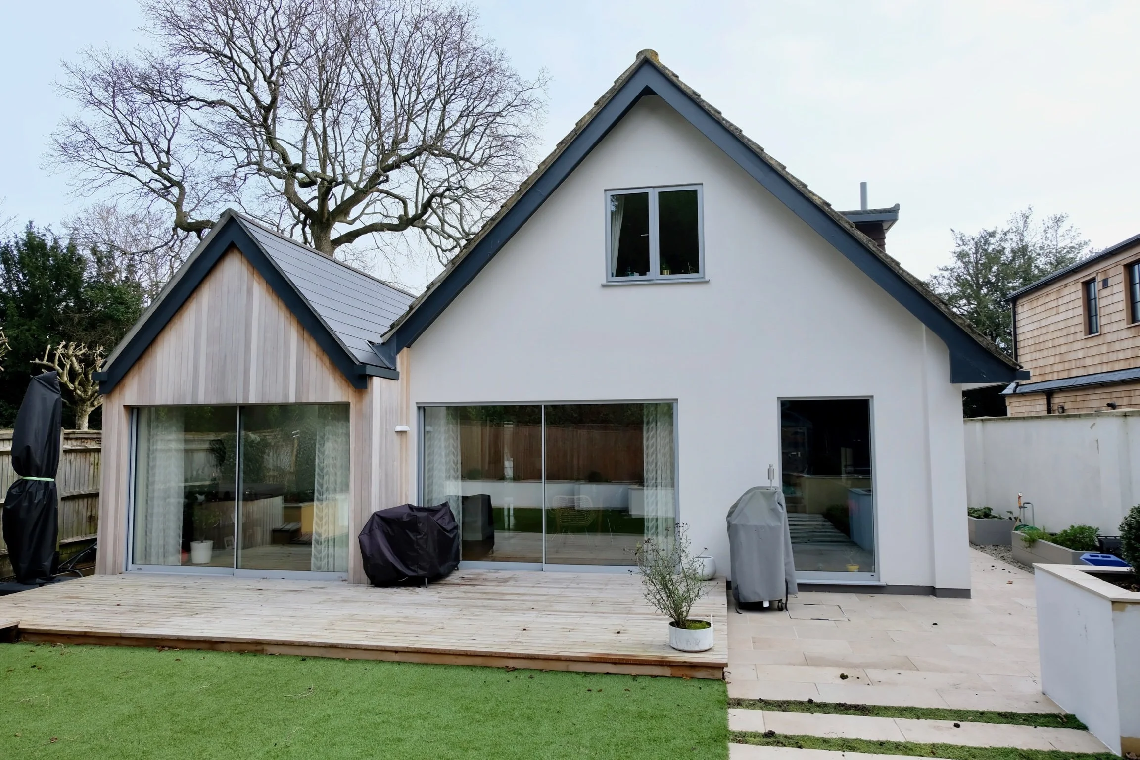 Modern house with large glass sliding doors, a wooden deck, and a small lawn area, backyard with trees and neighboring houses