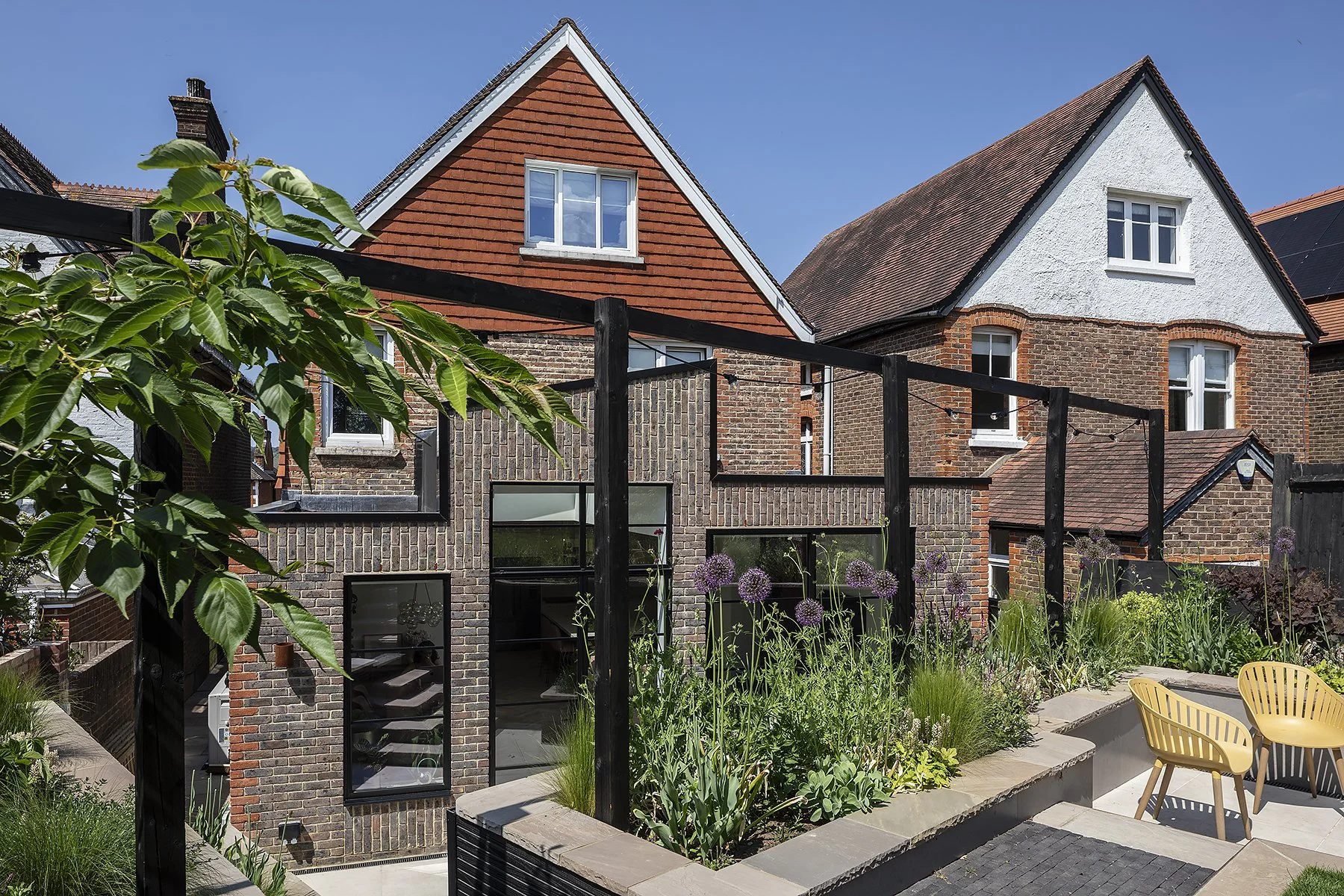 The image shows a backyard garden with patio furniture, plants, and a modern multi-story brick house with a gabled roof and multiple windows.