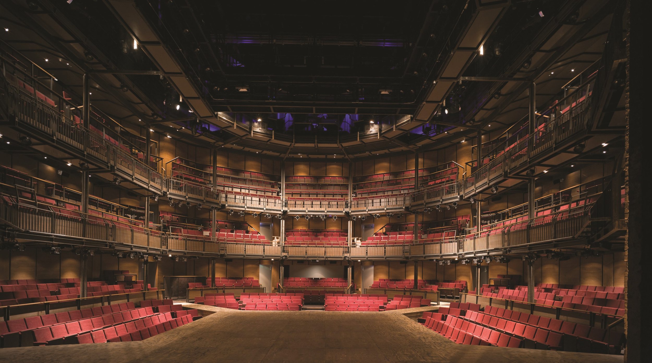 Theatre Auditorium with multiple tiers of red seats and a wooden stage in the foreground.
