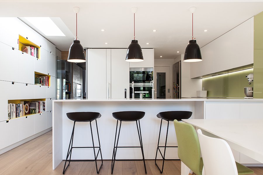 Modern kitchen with white cabinets, black pendant lights, black bar stools at a white island, and a dining area with green and white chairs.