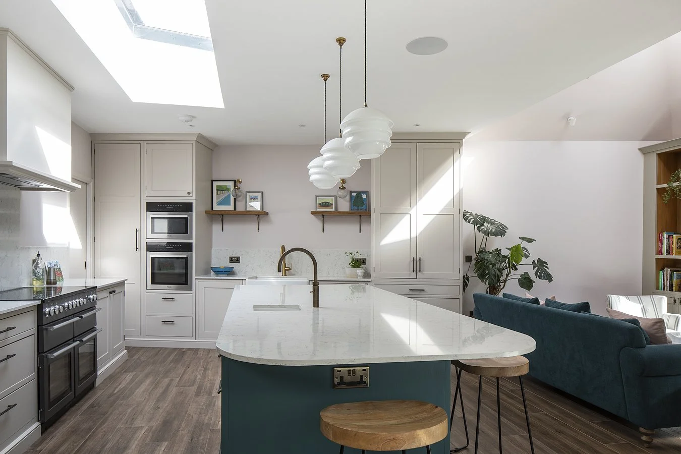 Modern kitchen with white cabinetry, a marble island with a gold faucet, gray wood flooring, hanging white pendant lights, and a living area with a teal couch and a large plant.
