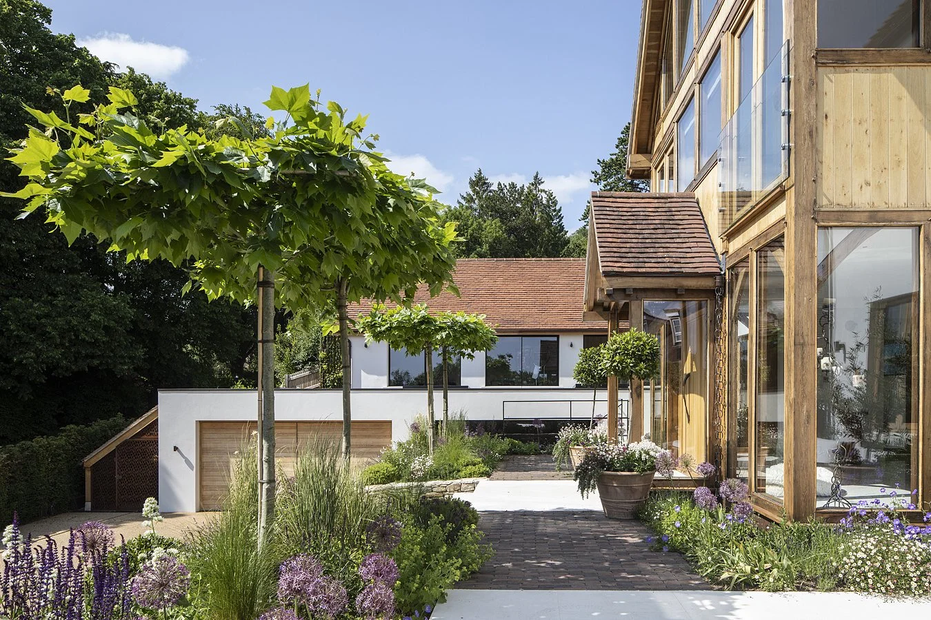 Modern house with large glass windows, wooden exterior, and a landscaped garden with trees, flowers, and brick pathways.