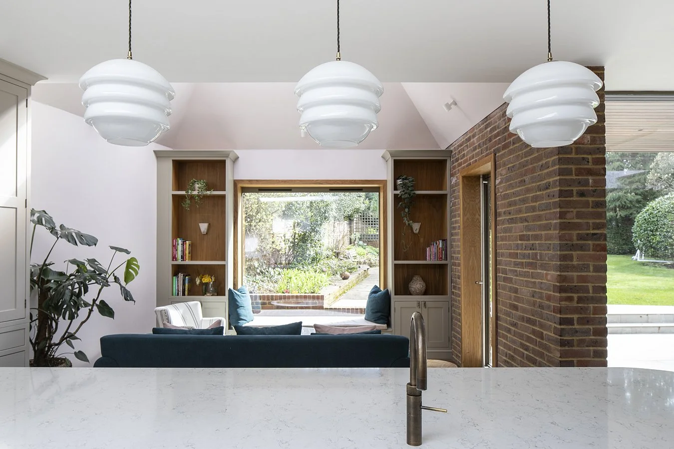 Modern kitchen with a white countertop, three hanging pendant lights, a brick wall, built-in bookshelves, and a window seat overlooking a garden.
