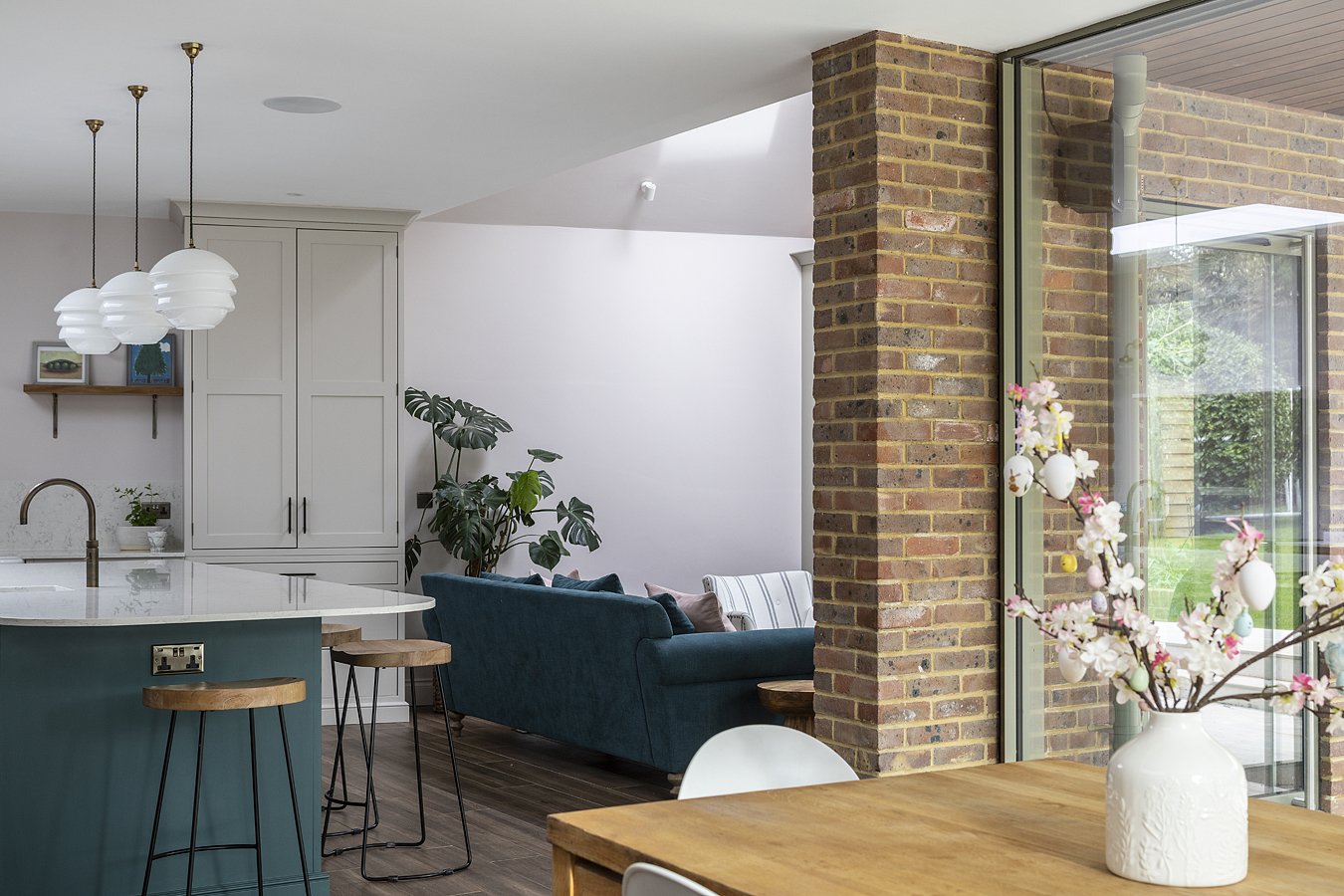 Interior view of a modern living space with a kitchen featuring a white countertop, pendant lights, and white cabinetry, a blue sofa, indoor plants, a brick wall, and a large window with a view of a green outdoor area, decorated with a vase of pink a