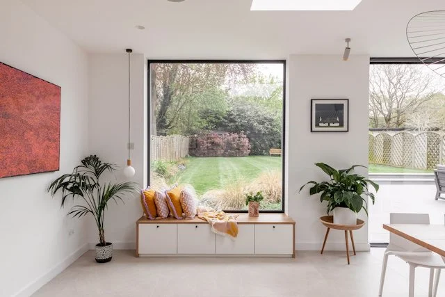 Bright modern living room with large window overlooking a backyard with green grass and trees, indoor plants, artwork on the wall, and a cozy window seat with colorful cushions.