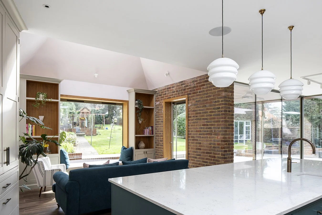 Living room with brick wall, large windows showing backyard with swing set, and modern white pendant lights over a white kitchen island