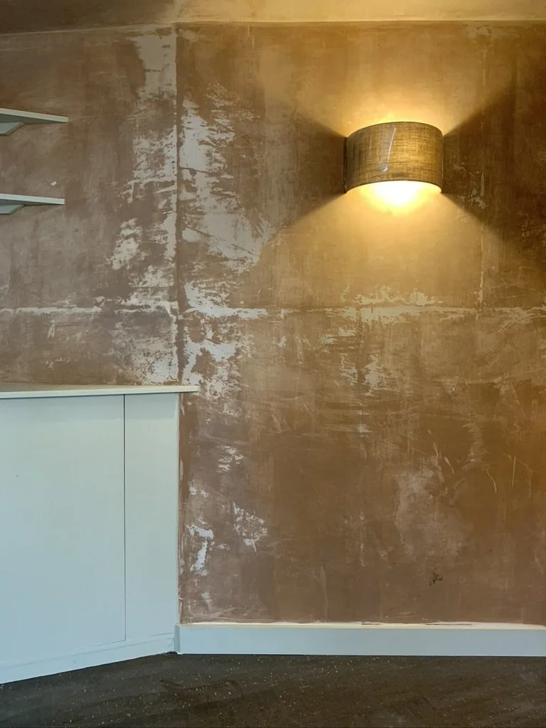 Interior wall with textured, distressed finish, illuminated by a wall-mounted light fixture with fabric shade, next to a white cabinet or counter, and dark flooring.