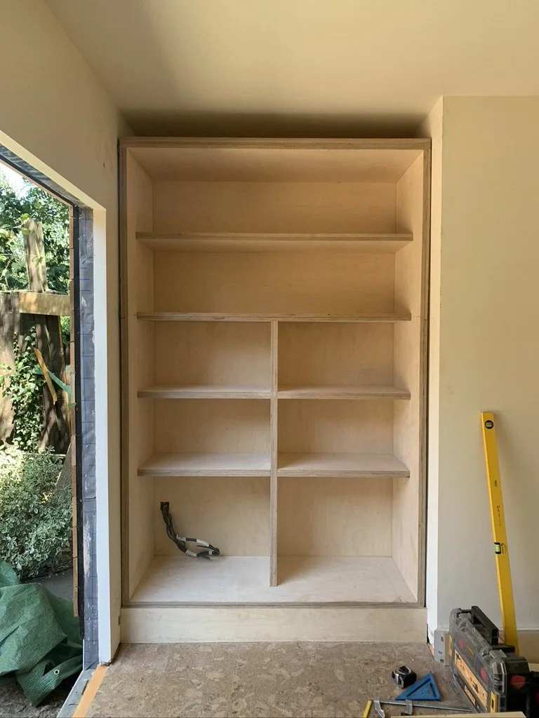 Empty built-in wooden shelving unit with five shelves and a lower section with electrical wires, situated in a room under construction next to an open door leading outside, with tools and a leveling tool placed on the floor.